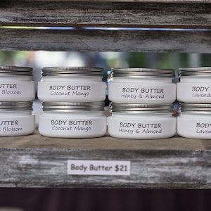 May include: A display of eight white jars of body butter with silver lids, arranged on a wooden shelf. Each jar is labeled with a different scent: Baja Cactus Blossom, Coconut Mango, Honey & Almond, and Lavender + Sage. The jars are neatly stacked in pairs.