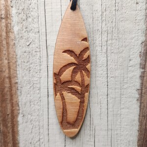 May include: A wooden pendant in the shape of a surfboard, featuring an engraved palm tree design. The pendant is light brown and hangs from a black cord. The background is a weathered, white-painted wooden surface.
