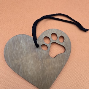May include: A wooden heart-shaped ornament with a paw print cutout and a black string for hanging. The ornament has a natural wood grain finish and is set against an orange background. A pet-themed decorative item.