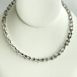 May include: A silver-toned necklace with a repeating geometric link design. The necklace is displayed on a white bust, showcasing its shape and style. The links are rectangular with a decorative element.