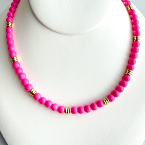May include: A vibrant pink beaded necklace with gold-colored accents. The necklace features small, round pink beads strung together, interspersed with cylindrical gold-colored spacers. The necklace is displayed on a white bust.