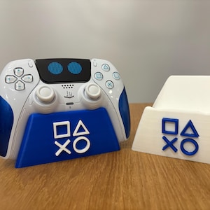 May include: A white and blue PlayStation 5 controller sits on a blue stand with the PlayStation logo. The stand is on a wooden surface. Next to the controller is a white stand with the PlayStation logo.