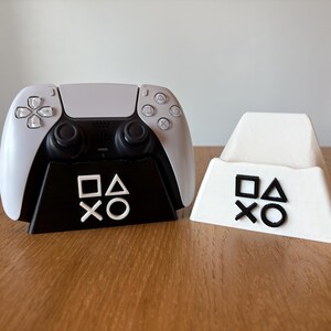 May include: A white PlayStation 5 controller sits on a black stand with the PlayStation symbols. A white stand with the PlayStation symbols is next to it.