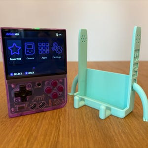 May include: A purple handheld gaming console with a screen displaying the text "onionOS" and icons for Favorites, Games, Apps, and Settings. The console has a directional pad, four buttons, and a select and back button. A light blue plastic stand with legs is next to the console.