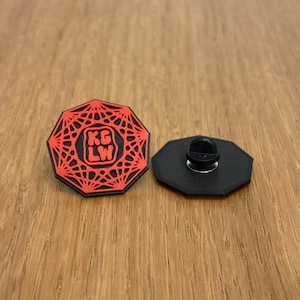 May include: A red and black enamel pin with a geometric design and the letters "KG LW" in the center. The pin is octagonal and has a black backing with a pin clasp.