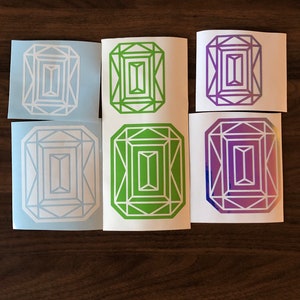 May include: Six emerald-shaped stickers in different colors. The stickers are on a wooden surface. The stickers are white, light blue, green, purple, and a gradient of purple and white.
