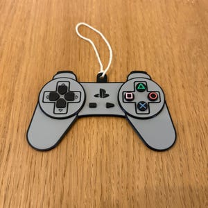 May include: A gray PlayStation controller ornament with a white string for hanging. The controller features black buttons and directional pad, and colored action buttons: green triangle, red circle, blue X, and a square.