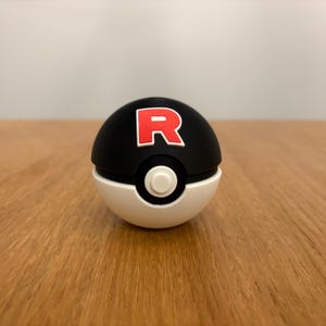 May include: A black and white novelty ball with a red "R" logo. The top half is black, the bottom half is white, and a white button is in the center. The ball is sitting on a wooden surface.