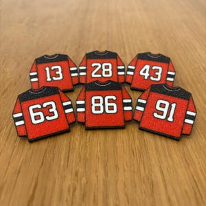May include: Six red and black jersey-shaped embellishments with white numbers: 13, 28, 43, 63, 86, and 91. The jerseys have black collars and sleeves with white stripes. The background is a light brown wooden surface.