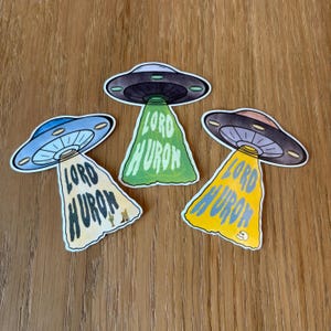 May include: Three stickers featuring UFOs with the text "LORD HURON" in a stylized font. The UFOs have a classic flying saucer design with a central light beam. The text is in green, yellow, and white.