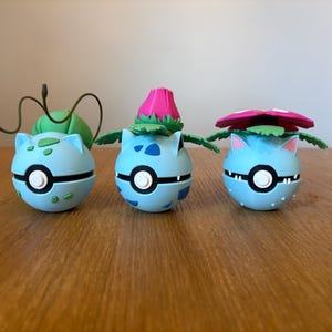 May include: Three light blue, spherical figurines with black and white accents, resembling a Poké Ball. Each has a different plant-like feature on top: a green bulb with tendrils, a pink flower bud, and a pink and green flower. They are displayed on a wooden surface.