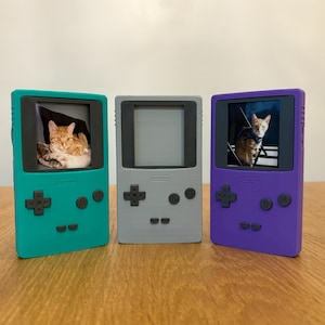 May include: Three colorful handheld gaming device replicas. The teal, gray, and purple devices feature a screen displaying a cat. The teal device shows an orange cat resting. The purple device shows a cat wearing a harness. The gray device has a blank screen.