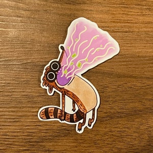 May include: A cartoon raccoon with large eyes and a purple tongue is blowing a stream of pink and green goo. The raccoon is brown and has a striped tail.