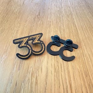 May include: Two black enamel pins with the number "33" in a stylized font. One pin has a gold outline and fill, the other is a solid black shape with two pin backs. The pins are on a wooden surface.