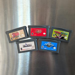 May include: Five different Game Boy Advance game cartridges. The cartridges are black with colorful labels. The games are "The Legend of Zelda: The Minish Cap", "Final Fantasy IV", "Metroid Fusion", "Mario & Luigi: Superstar Saga", and "The Legend of Zelda: Four Swords".