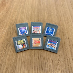 May include: Six gray Game Boy cartridges with colorful game covers. The cartridges are arranged in two rows of three. The games are Tetris, Zelda, Super Mario Land, Kirby's Dream Land, Final Fantasy, and a game with a red, white, and blue cover.