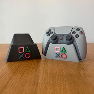 May include: A gray PlayStation 5 controller sits on a gray stand with the PlayStation symbols in pink, green, and blue. The controller is on a wooden surface. A black stand with the PlayStation symbols in pink, green, and blue is also on the surface.