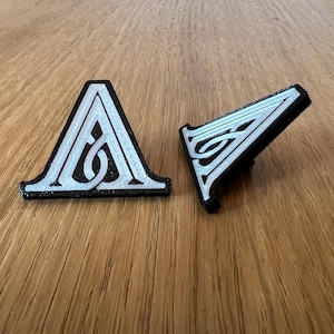 May include: Two black triangular-shaped pins with a white design of the letter "A" with a swirl inside. The pins are on a wooden surface.