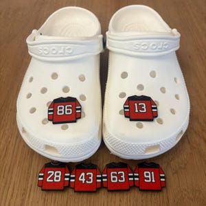 May include: Pair of white Crocs clogs with black and red jersey-style charms. One clog features the number "86" and the other "13". Additional charms with the numbers "28", "43", "63", and "91" are also present.