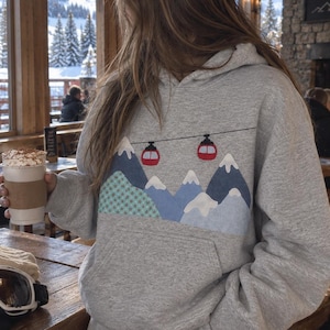 Op de afbeelding: Een grijs gemêleerde hoodie met een grafische print van blauwe bergen met witte besneeuwde toppen en twee rode kabelbaanwagentjes aan een zwarte lijn. De trui is voorzien van een kangoeroezak en lange mouwen met geribbelde boorden.