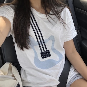 May include: A white short-sleeved t-shirt featuring a light blue graphic outline of an electric guitar body. Three vertical black stripes with white polka dots extend from the shoulder down to a black rectangular bridge with white polka dots.