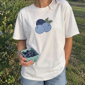 May include: A white short-sleeve t-shirt featuring a central applique design of three blueberries in shades of dark and light blue with two green leaves, accented by dark blue stitched outlines and small cross-stitch details on each berry.
