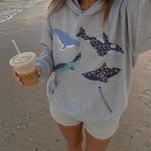 Könnte beinhalten: Ein graumelierter Kapuzenpullover mit vier Stoffapplikationen von Meerestieren, darunter ein hellblauer Wal, ein schwarz-weiß gemusterter Orca, ein blaugrün gemusterter Hai und ein floral gemusterter Stachelrochen, mit einem sichtbaren Nahtdetail.