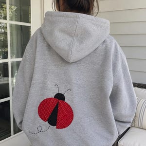Op de afbeelding: Een lichtgrijze hoodie met een grote applicatie van een lieveheersbeestje op de achterkant. Het lieveheersbeestje heeft een zwarte kop en twee rode vleugels met kleine witte stippen. Een zwarte stippellijn onder het insect suggereert een vlucht.