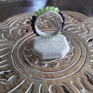 May include: A beaded bracelet featuring alternating rows of green and black beads. The bracelet is displayed on a gray, hexagonal jewelry display stand. The background is a carved wooden surface.