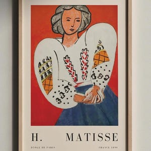 Peut inclure: Une impression d'art encadrée représentant une illustration d'Henri Matisse d'une personne en chemisier blanc aux manches à motifs sur fond rouge vif. Le texte H. MATISSE, ECOLE DE PARIS et FRANCE 1890 est imprimé en noir en bas.