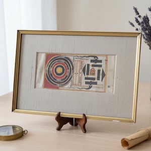 May include: Framed artwork with a gold-colored frame, featuring a detailed, colorful illustration. The artwork is displayed on a wooden stand. The illustration has concentric circles and geometric shapes, with red, black, and yellow hues. A compass and a rolled scroll are also visible.