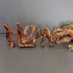 Può includere: Un'insegna rustica in legno con la parola "HOME". Le lettere sono realizzate in legno naturale e strutturato, che mostra diverse tonalità di marrone e beige. L'insegna è un elemento decorativo, perfetto per aggiungere un tocco di natura a qualsiasi casa.