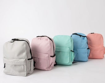 May include: Five different colored backpacks lined up in a row. The backpacks are all the same style, with a single zippered pocket on the front and two adjustable straps. The colors are gray, pink, light green, light blue, and peach.