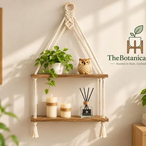 May include: A two-tier wall-mounted macrame shelf featuring natural cream-coloured cotton rope, two dark wood planks, a green potted plant, a small owl figurine, two pillar candles, and a reed diffuser with the text TheBotanicalDecor.