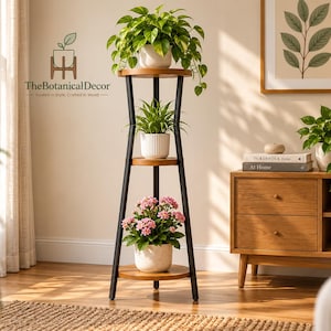 May include: A three-tier plant stand with a black metal frame and circular wooden shelves. The stand holds a trailing green plant on top, a small green plant in a white ribbed pot in the middle, and a pink flowering plant in a white pot at the base.