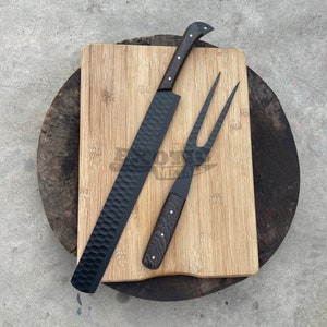 Puede incluir: Un juego de cuchillo y tenedor para trinchar negros con mangos de madera descansa sobre una tabla de cortar de bambú. El cuchillo tiene una hoja texturizada y el tenedor tiene dos púas. El juego se exhibe sobre una superficie de madera.