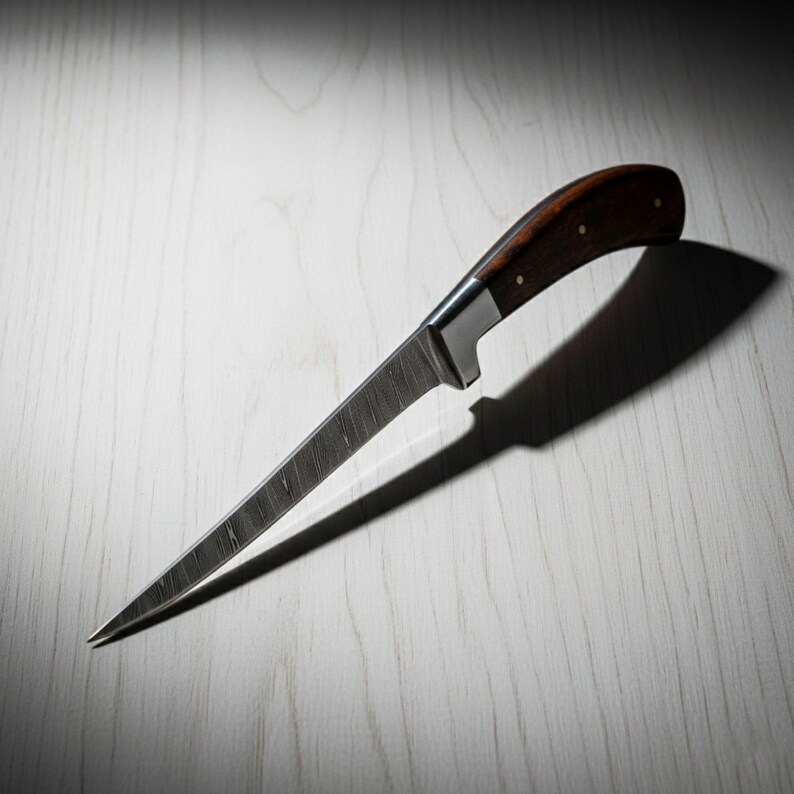 May include: A long, slender knife with a dark wood handle and a textured, silver blade. The handle is curved and secured with small rivets. The knife is angled, casting a shadow on the light-colored surface.
