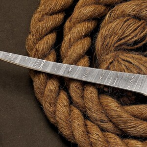 May include: A Damascus steel knife with a textured blade and a white handle. The knife is set against a backdrop of coiled brown rope, creating a rustic aesthetic. The blade's pattern is a prominent feature.