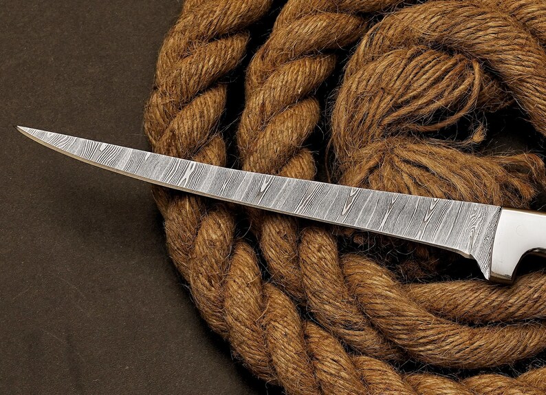 May include: A Damascus steel knife with a textured blade and a white handle. The knife is set against a backdrop of coiled brown rope, creating a rustic aesthetic. The blade's pattern is a prominent feature.