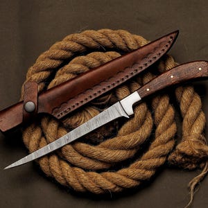 May include: A handcrafted knife with a patterned blade and a wooden handle, resting on a coil of rope. The knife is accompanied by a brown leather sheath. The blade is approximately 8 inches long.