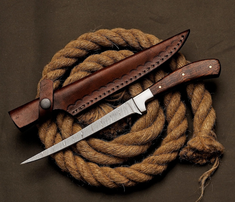 May include: A handcrafted knife with a patterned blade and a wooden handle, resting on a coil of rope. The knife is accompanied by a brown leather sheath. The blade is approximately 8 inches long.