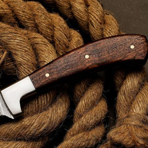 May include: Close-up of a handcrafted knife with a dark wood handle and a silver blade. The handle is secured with brass rivets. The knife rests on a thick, brown rope, highlighting its rustic aesthetic.