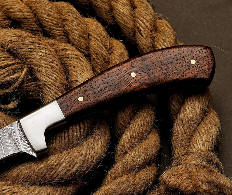 May include: Close-up of a handcrafted knife with a dark wood handle and a silver blade. The handle is secured with brass rivets. The knife rests on a thick, brown rope, highlighting its rustic aesthetic.