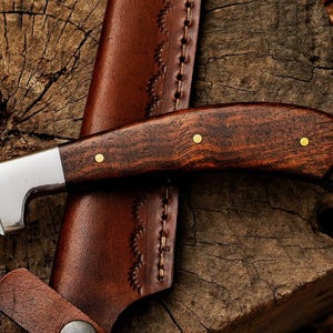 May include: A close-up of a hunting knife with a dark wood handle and a silver blade. The handle has brass rivets. The knife rests in a brown leather sheath with decorative stitching. The background is a weathered wooden surface.