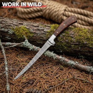 May include: A hunting knife with a patterned steel blade and a dark wood handle. The handle is secured with brass rivets. The knife rests on a bed of pine needles and a moss-covered log. The text "WORK IN WILD" is in the upper left corner.