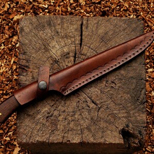 May include: A handcrafted knife with a dark wooden handle and a brown leather sheath. The sheath is stitched along the edges and secured with a metal button. The knife rests on a weathered wooden block.