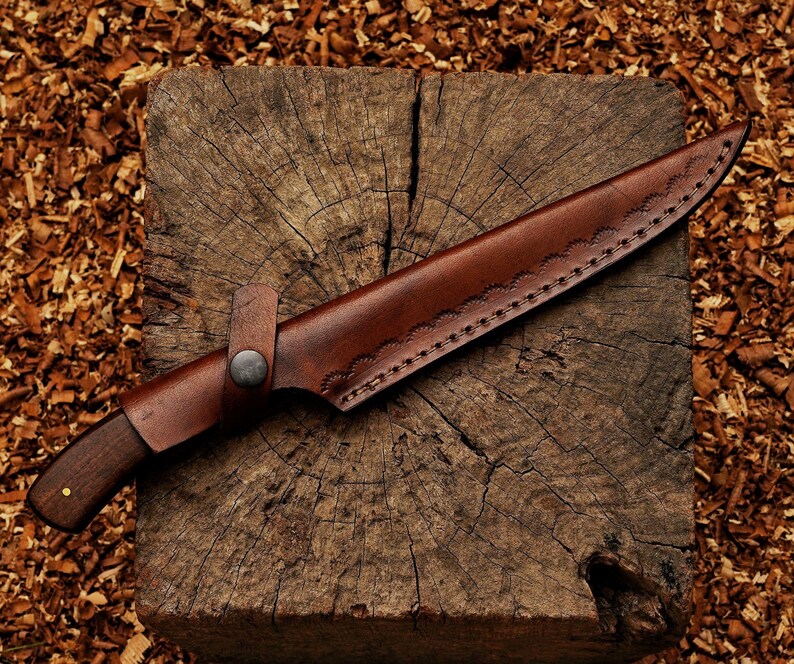 May include: A handcrafted knife with a dark wooden handle and a brown leather sheath. The sheath is stitched along the edges and secured with a metal button. The knife rests on a weathered wooden block.
