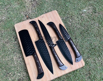 May include: A set of five black kitchen knives with wooden handles, displayed on a light brown wooden cutting board. The blades have a textured surface. The knives are arranged on a bed of green grass.