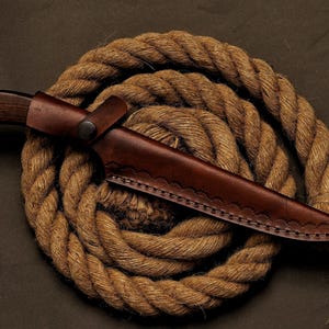 May include: A hunting knife with a dark wooden handle and a brown leather sheath. The knife is resting on a coiled rope, creating a rustic and outdoorsy aesthetic. The sheath has decorative stitching.