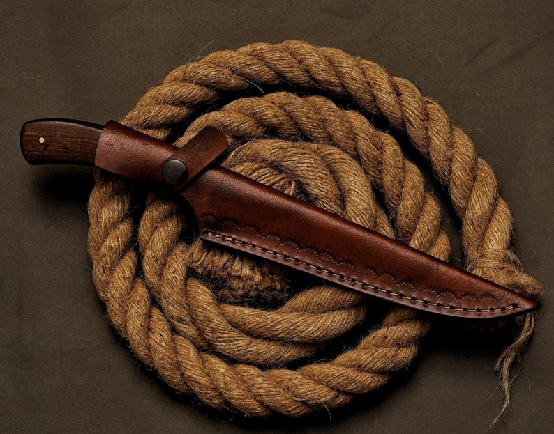 May include: A hunting knife with a dark wooden handle and a brown leather sheath. The knife is resting on a coiled rope, creating a rustic and outdoorsy aesthetic. The sheath has decorative stitching.